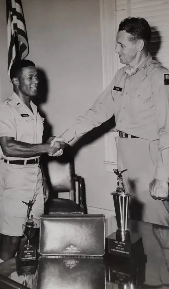Schwarzweißfoto: Johnson in Uniform gibt einem älteren Offizier die Hand, auf dem Tisch zwei Pokale für das 3-Meter-Kunstspringen in Fort Totten