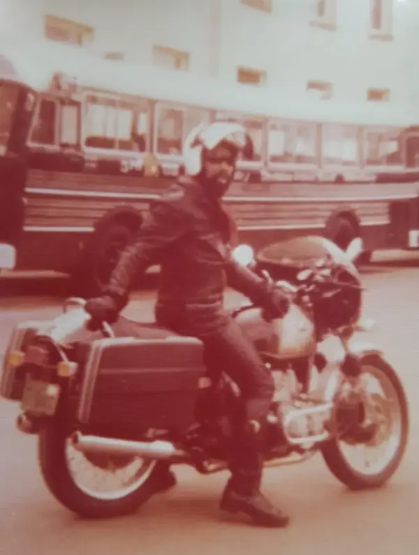 Farbfoto aus den 1970er Jahren: Johnson mit Bart, Lederjacke, Helm und Sonnenbrille auf einem Tourenmotorrad mit Koffern, im Hintergrund ein Stadtbus